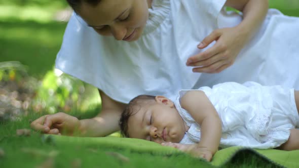 Mother Kisses Her Baby Sleeping on a Green Grass Outdoors. Happy Smiling Young Mother and Child in