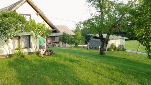 Flying between fruit trees following man lawn mowing grass with small lawnmower. Aerial 4k view.