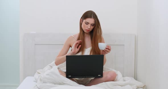 Young Woman Freelancer Is Sitting on the Bed Drinking Coffee and Working on a Laptop, Morning Time