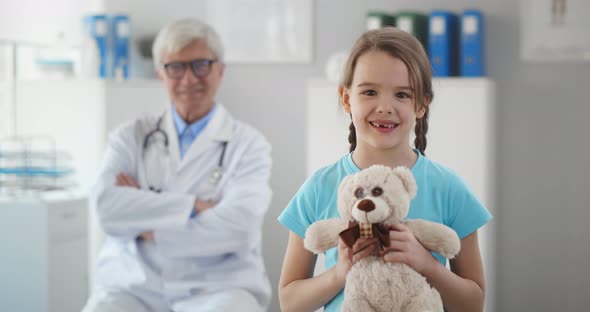 Smiling Healthy Little Girl Hugging Teddy Bear in Doctor Office Looking at Camera