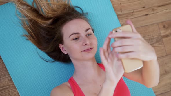 Fitness Healthy Woman in Sportswear Is Lying on the Mat and Holding a Smartphone in Her Hands on