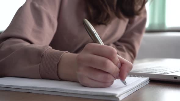 A Girl Takes Notes in a Notebook From Her Laptop