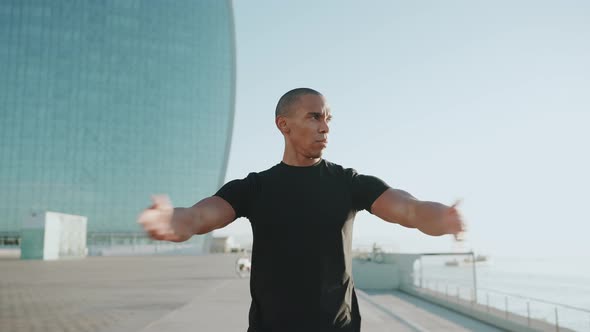 Concentrated bald sportsman wearing black t-shirt does hands warm-up outdoors