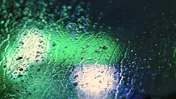 Close up of rain water drops on car window glass with blurred traffic lights background at night.