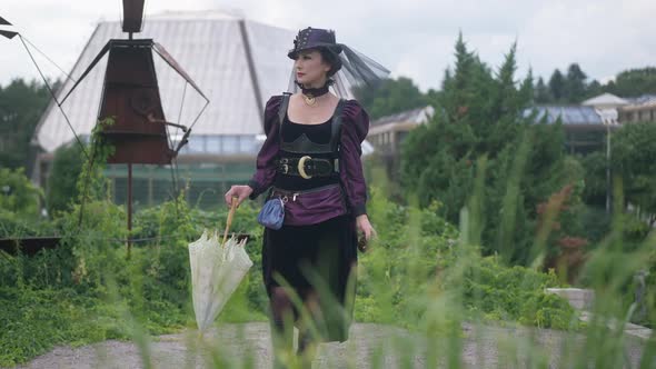 Confident Slim Woman in Purple Steampunk Dress and Hat Walking with Sun Umbrella and Gun Leaving