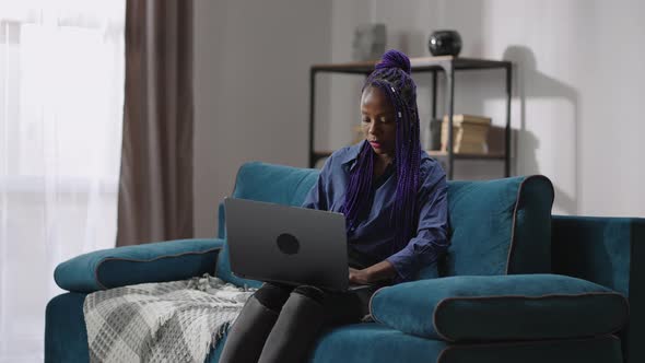 Young Black Woman with Purple Dreadlocks is Working Remotely with Laptop at Home at Weekend