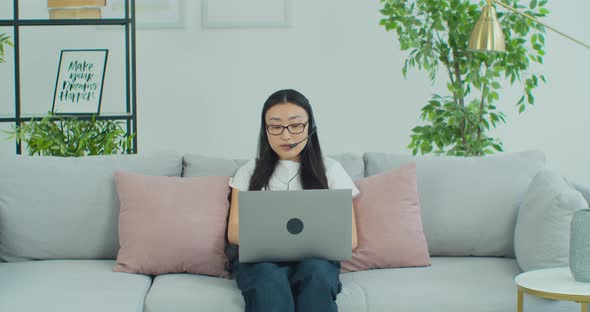 Asian Woman in Glasses Sit on Couch Wear Headset Use Laptop Having Distant Communication