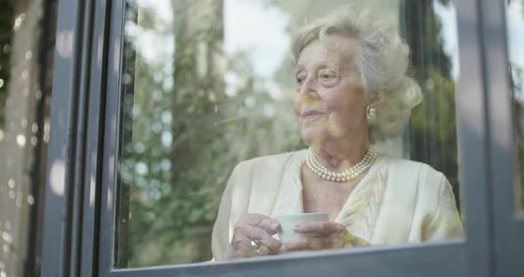 Senior Grandma Woman Enjoying Drinking Cup of Tea or Coffee Near Window Looking Outside