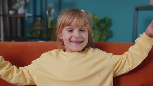 Smiling Child Girl Kid on Home Sofa Looking at Camera Makes Heart Gesture Demonstrates Love Sign