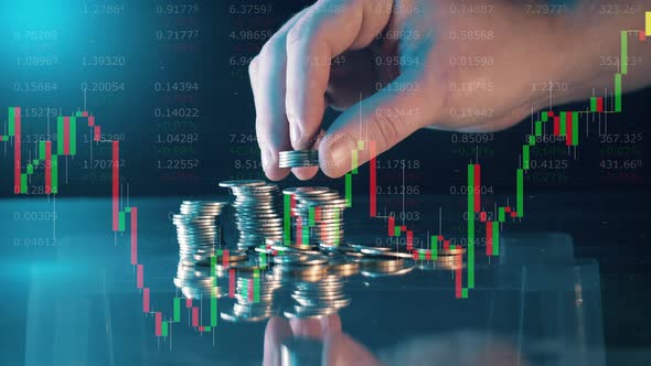 A Hand Adds Coins to a Pile with Stock Charts on the Screen