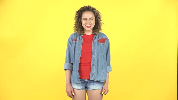 Lovely Curly Girl Smiles Broadly and Winks on Yellow Background at Studio