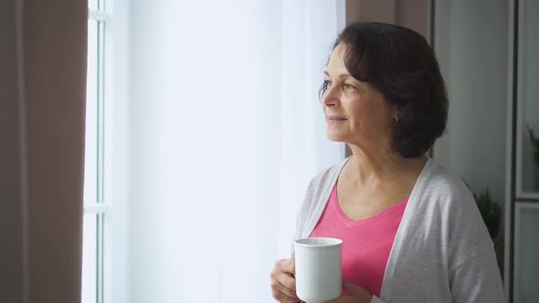 American Elderly Woman Drinking Coffee and Watching Window in Home Room