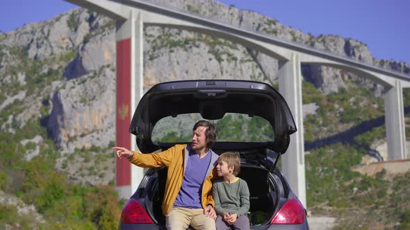 A Man and His Son are Sitting and Chatting in Their Car with the Moracica Bridge in the Background