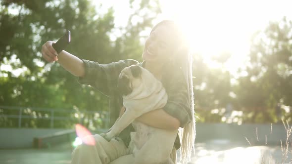 Smiling Girl Taking Selfie Photo with Cute Pug Puppy in Green City Park Holding Smartphone