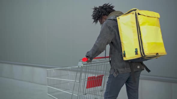 Young African American Courier with Yellow Food Delivery Bag Pushing Shopping Cart in Urban City