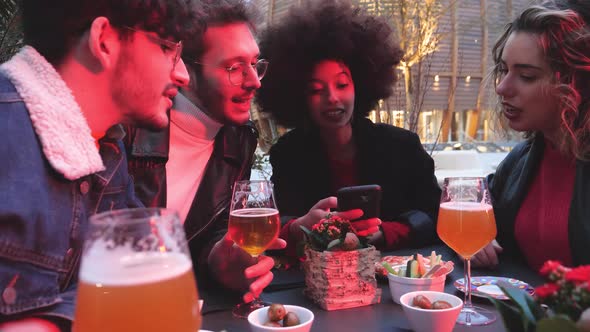 Group of four multiethnic friends sitting outdoor clubbing using smartphone