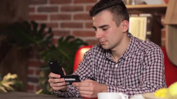 Handsome Man Making Purchase Online, Typing Credit Card Details on Black Smartphone in the Modern