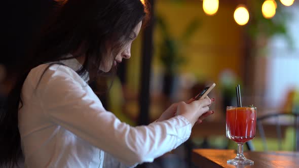 Pretty young dark-haired girl is sitting in a cafe and taking a photo of her cocktail for Instagram.