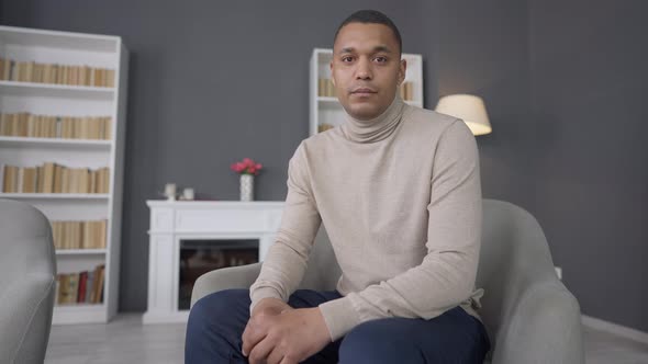 Confident Young African American Man Sitting on Comfortable Armchair at Home Looking at Camera