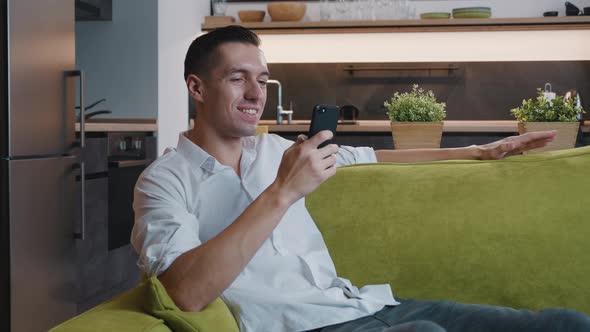 Smiling Young Man Using Smartphone While Relax on Sofa at Home. Cheerful Male Taping While Texting