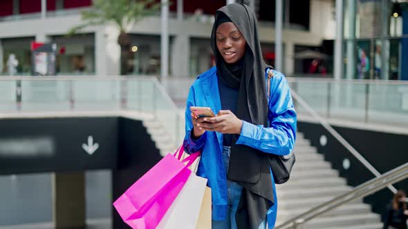 Black Shopaholic Adjusting Hijab and Using Smartphone