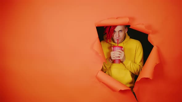 Young Man Drinking Through Soft Drink Straw in Hole of Orange Background