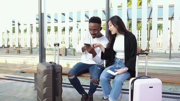 Mixed Race Friends Sitting on Tram or Bus Stop with Suitcases, Waiting for Transport