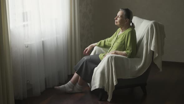 Senior Woman Sitting on Armchair and Looks Outside the Window
