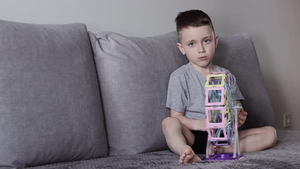 A boy in a gray T-shirt sits on a bed near a white wall and plays with a Ferris wheel