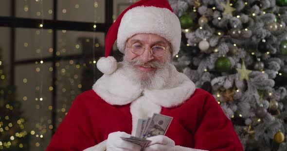 Cheerful Elderly Man Dressed Like Santa Claus Quickly Counts Money at Home Against the Background of