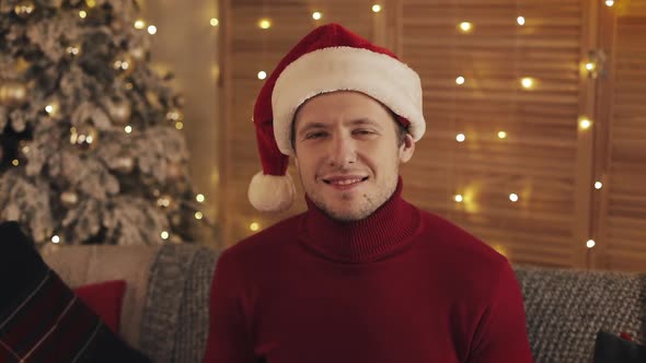 Portrait of Smiling Man in Santa Hat Looking at the Camera on Christmas Tree Background