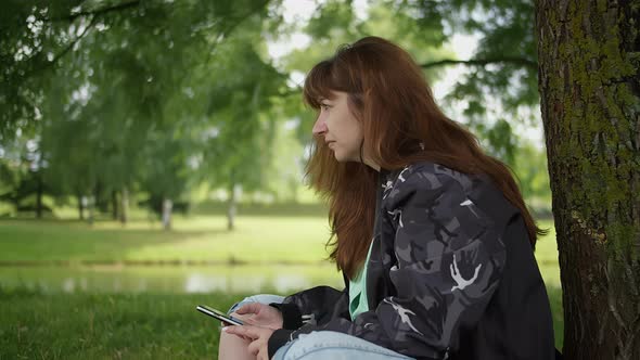 Woman Sitting Under a Tree in the Park Uses the Phone and Thinks