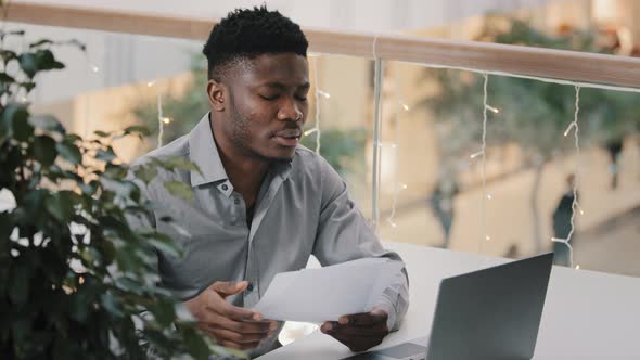Concentrated Frustrated Upset African American Man Sitting at Desk with Laptop Looking at Documents