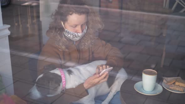 Woman Wearing Mask Using Smartphone In Cafe With Dog During Coronavirus Pandemic.