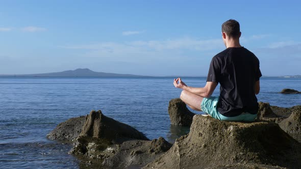 Young fit man meditating in yoga pose on rocks at the beach in the morning