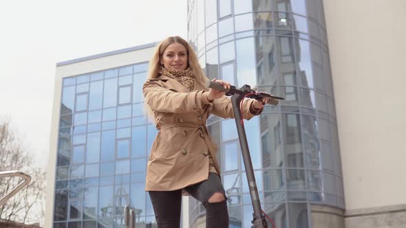 Active Young Stylish Blonde Girl in a Beige Trench Coat and Jeans Stands with an Electric Scooter on