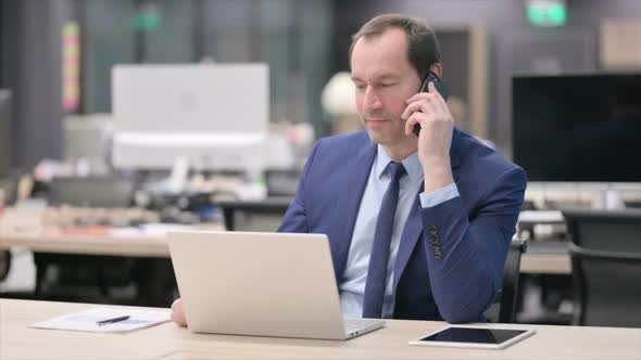 Businessman Talking on Smartphone While Using Laptop in Office