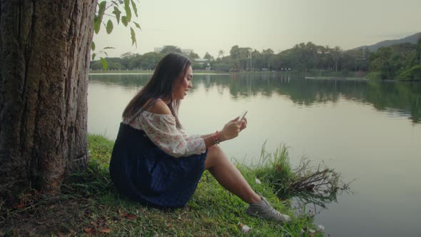 Transgender Asian Woman Sitting By a Tree in the Park Texting on Her Phone