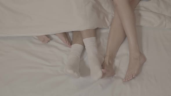 Man legs in white socks touched by two women bare feet laying on bed under blanket white bed sheets