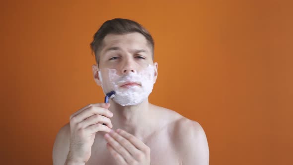 Portrait of a Beautiful Young Man Shaving His Face Using Foam and Razor, Isolated Orange Background