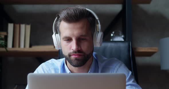 A Focused Young Man with Headphones is Typing on His Laptop Computer Sitting at Home