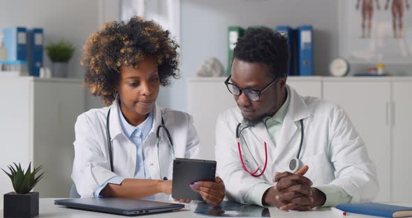 African Doctors Discussing Patient Medical Test Results on Tablet Sitting at Desk in Clinic Office