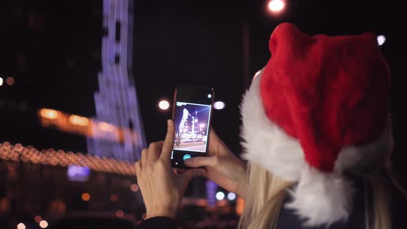 A woman in a Santa Claus hat takes pictures of night city with her smartphone