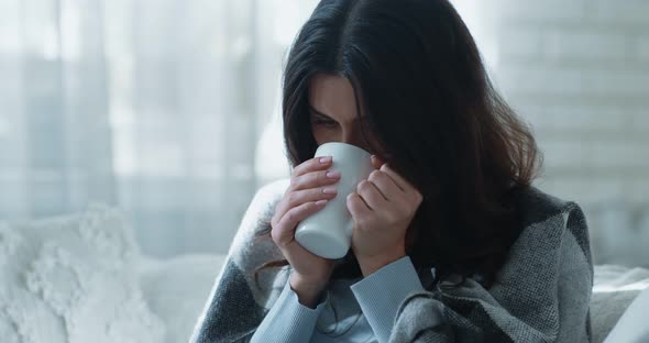 Young Depressed Woman Warming Hands of Cup, Enjoying Hot Tea, Sitting on Sofa Alone Wrapped in Plaid