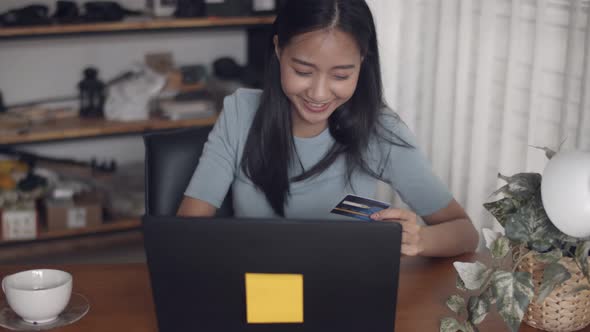 Attractive portrait young Asian female holding credit card using laptop shopping online at home.