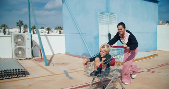 Young Happy Mother Running with Shopping Cart with Kid Sitting in It in Parking