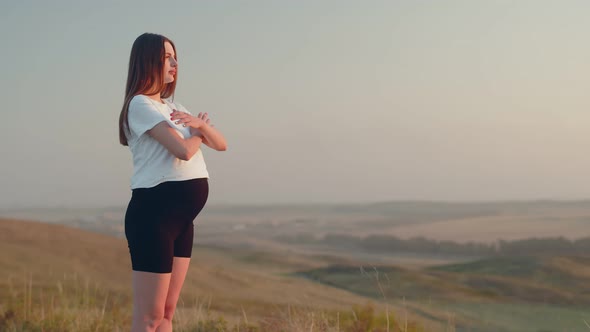 Caucasian Pregnant Woman in Sports Tshirt and Shorts Stands Outdoor and Folds Arms on Chest Then