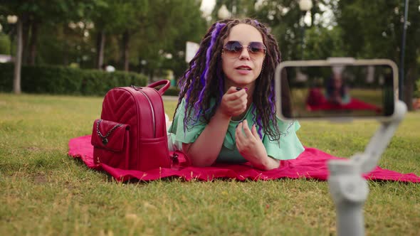 A Woman with a Stylish Hairstyle Lies on a Blanket in the Park and Records a Video Blog Using Her