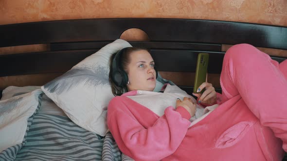 Girl in Pajamas on a Bed in Headphones is Talking on a Video Call on the Phone