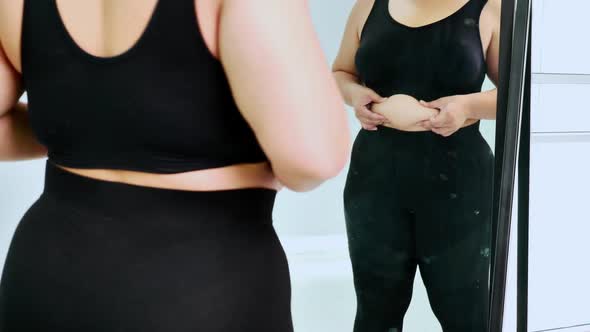 Overweight young woman in sportswear looking her belly fat into the mirror